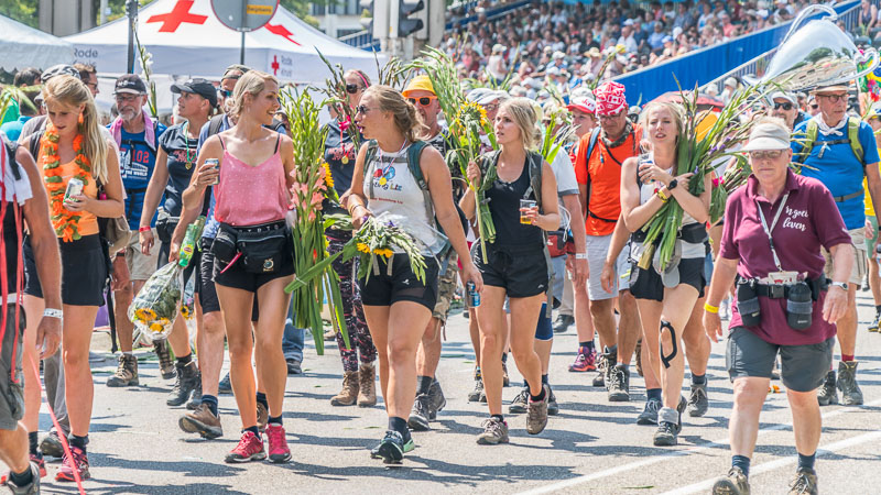 Numerous participants march through a tree-lined avenue during a major walking event. A banner reading “4Daagse Rustpost” by KWBN hangs above them.