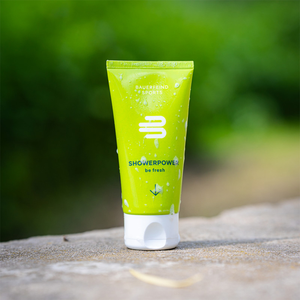 A green tube of Bauerfeind Sports "ShowerPower" shower gel stands on a rock outdoors, covered in water droplets. The tube features the slogan "be fresh" and gives a refreshing vibe.