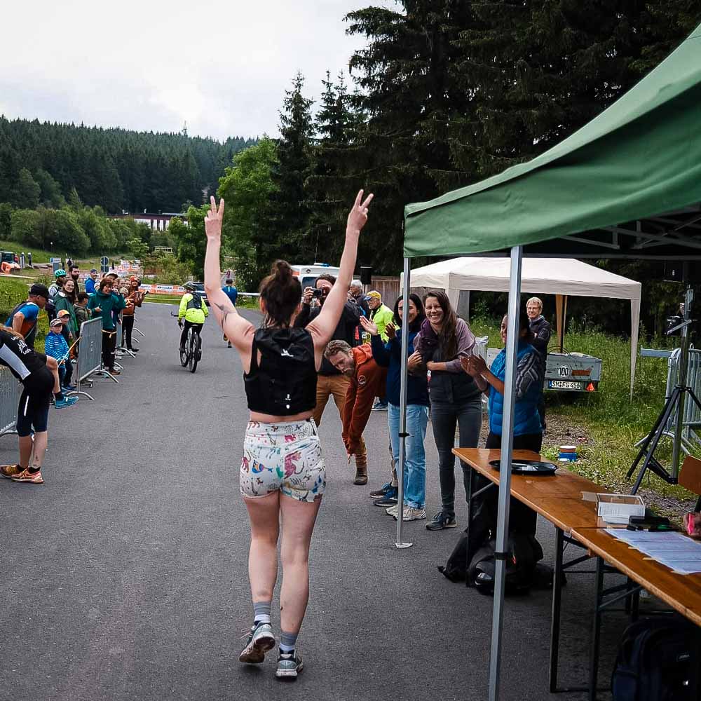 A woman in sportswear jogs through the Rennsteig and raises both arms to make the peace sign while the spectators cheer her on