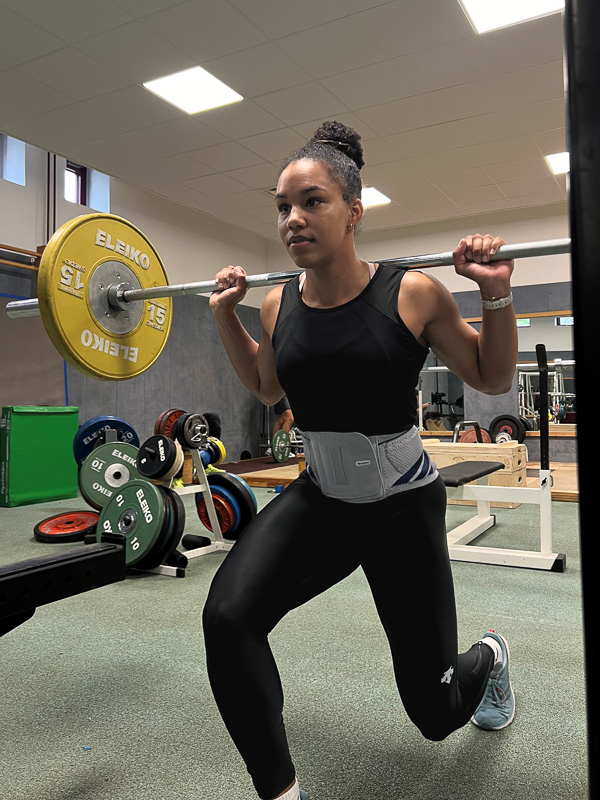 Deborah Levi macht im Fitnessstudio einen Ausfallschritt nach vorn. Auf ihren Schultern trägt sie eine Langhantel, um das Becken hat sie eine Bandage.