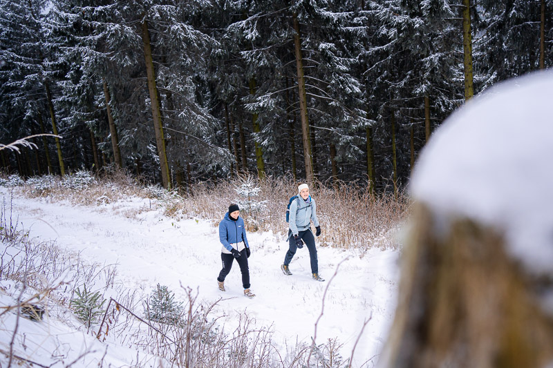 Zwei Frauen wandern durch eine verschneite Waldlandschaft. Sie tragen warme Winterkleidung und nutzen Wanderstöcke, während sie einen Pfad entlanglaufen. Im Hintergrund sind schneebedeckte Bäume und ein dichter Wald zu sehen.