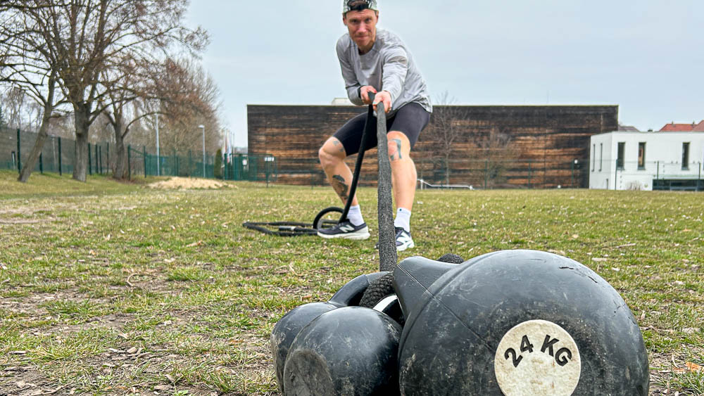 Athlete drags two kettlebells with a strap across a field during HYROX training outdoors – functional strength training without a gym.