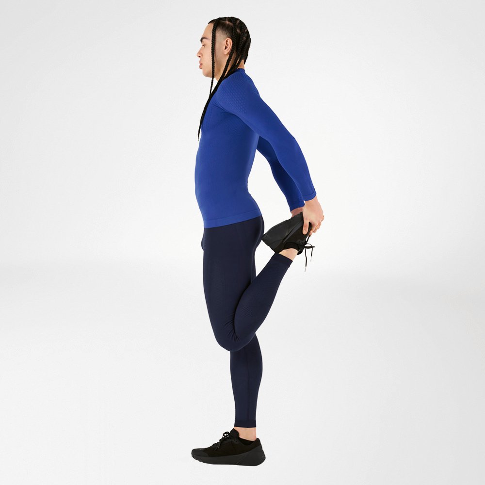 An athlete wearing a blue compression sports wear outfit, consisting of a long-sleeve shirt and tights, performs a stretching exercise, holding one leg back, against a white background. The outfit provides high comfort and optimal muscle support.