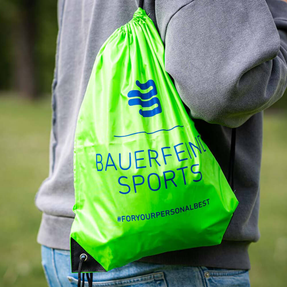 A person carries a green Bauerfeind Sports drawstring bag on their shoulder outdoors.