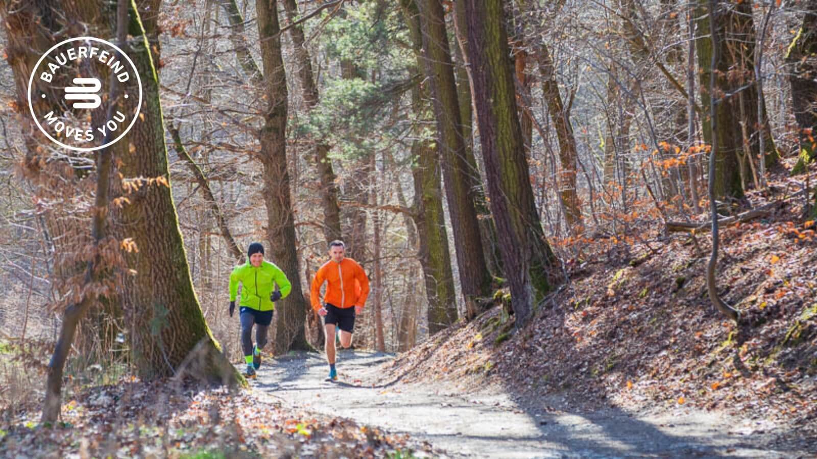 Two runners in brightly colored jackets (green and orange) jog along a sunlit forest path, surrounded by tall trees and dry leaves. Both are wearing distinctive socks that may indicate functional sportswear.