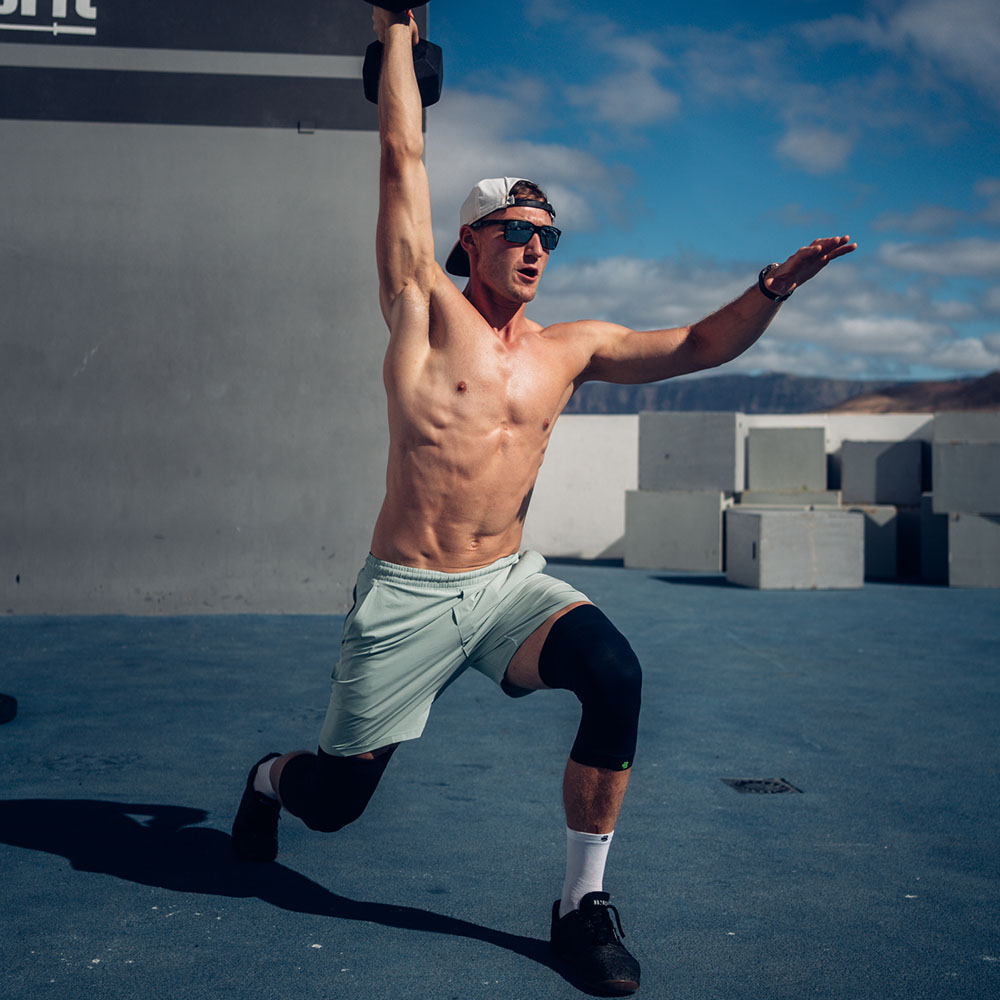 Maximum stability – even during intense workouts! A fit man performs a single-leg lunge with an overhead dumbbell while wearing a black knee brace.