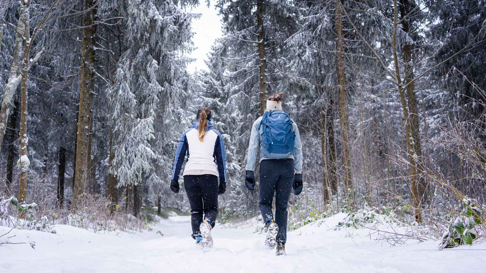 Zwei Frauen wandern durch eine verschneite Waldlandschaft. Sie tragen warme Winterkleidung und nutzen Wanderstöcke, während sie einen Pfad entlanglaufen. Im Hintergrund sind schneebedeckte Bäume und ein dichter Wald zu sehen.