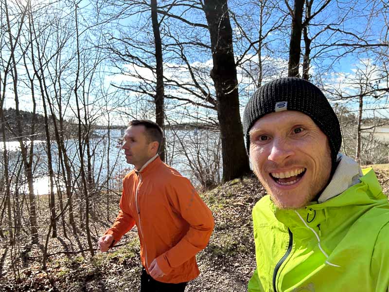 Two men are jogging along a sunny forest path beside a lake. One wears a neon green jacket and a beanie, the other wears an orange jacket.