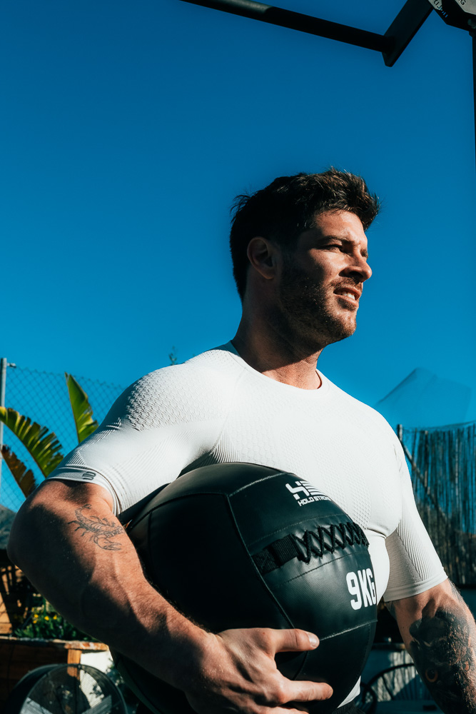 Athlete stands outdoors holding a black 9 kg medicine ball, wearing a fitted performance shirt and looking ahead in the sunlight – functional strength work in a hybrid athlete training setting.