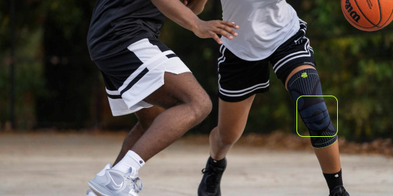Two basketball players in action on an outdoor court. The player in the foreground is wearing a black knee support and is dribbling while being closely guarded by the opponent. The movement conveys game intensity and athletic focus.