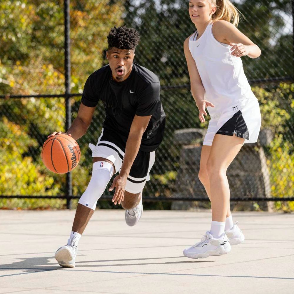 Two basketball players in a one-on-one duel on an outdoor court. The player in black wears a white compression knee sleeve and dribbles the ball while the opponent in white defends.