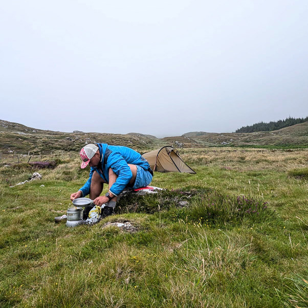Fitness-Challenge: Wenn die Natur zur Küche wird und jede Mahlzeit ein Abenteuer ist. Eine Person in blauer Jacke sitzt auf einer Wiese in hügeligem Gelände vor einem kleinen Zelt und bereitet eine Mahlzeit auf einem Campingkocher zu. Der Himmel ist bewölkt, und die Landschaft wirkt rau und naturbelassen.