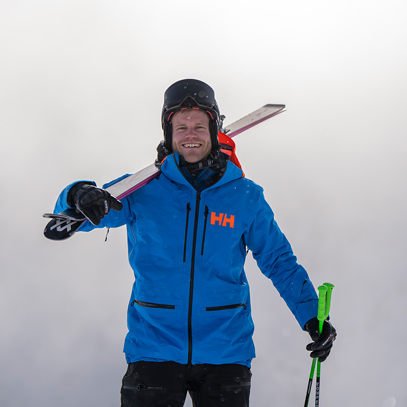 A man stands in front of a foggy background, wearing a blue jacket and a helmet with ski goggles and has his skis on his shoulder.