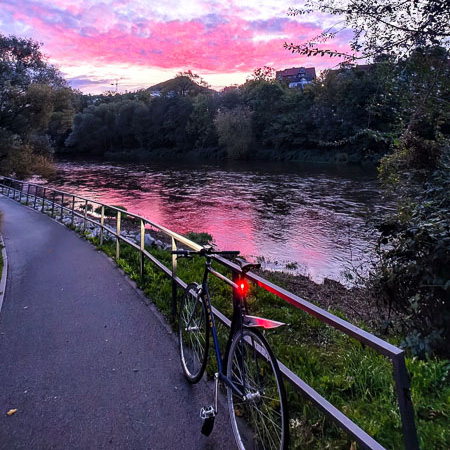 Sonnenuntergangszauber – wenn die Fitness-Route zur romantischen Kulisse wird. Ein Fahrrad lehnt an einem Geländer an einem Flussufer, während der Himmel in lila und rosa Tönen leuchtet. Im Hintergrund spiegeln sich die Farben des Himmels im ruhigen Wasser des Flusses.