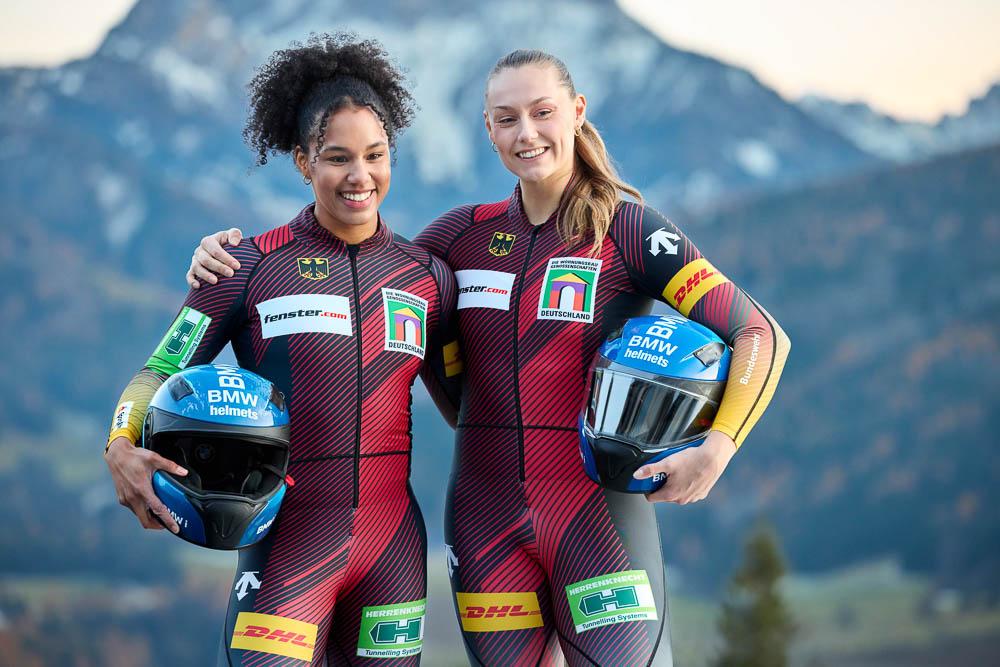 Two professional female athletes wearing German national bobsleigh suits stand side by side in front of an alpine mountain backdrop, holding their helmets and appearing confident and focused.