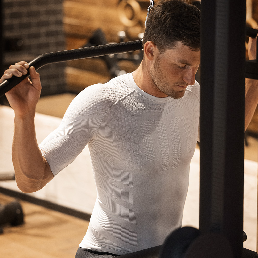 Athlete wearing a beige short-sleeve compression shirt while training on a strength machine at the gym. The seamless, tight fit provides muscle support and promotes stable posture.