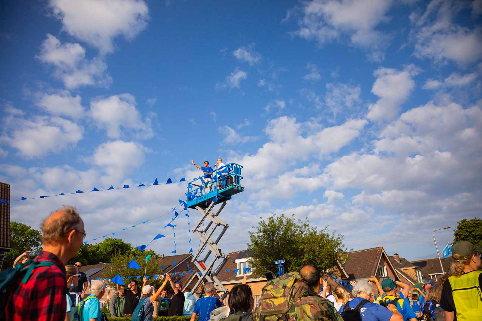 Two people cheer and wave from a lift platform at the 4Daagse in Nijmegen, while participants and spectators walk below blue flags.