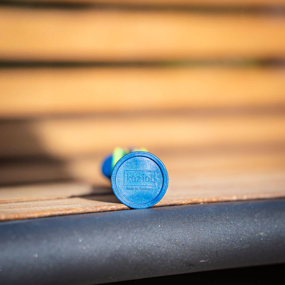 Close-up of the bottom of a blue piece of cutlery lying on a wooden bench. The bottom features the logo "Koziol Made in Germany."