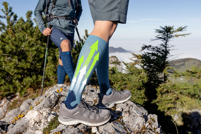 Two hikers on a rocky mountain trail wearing compression socks with a dynamic design featuring green and white arrows. The socks highlight support and energy flow during the activity.