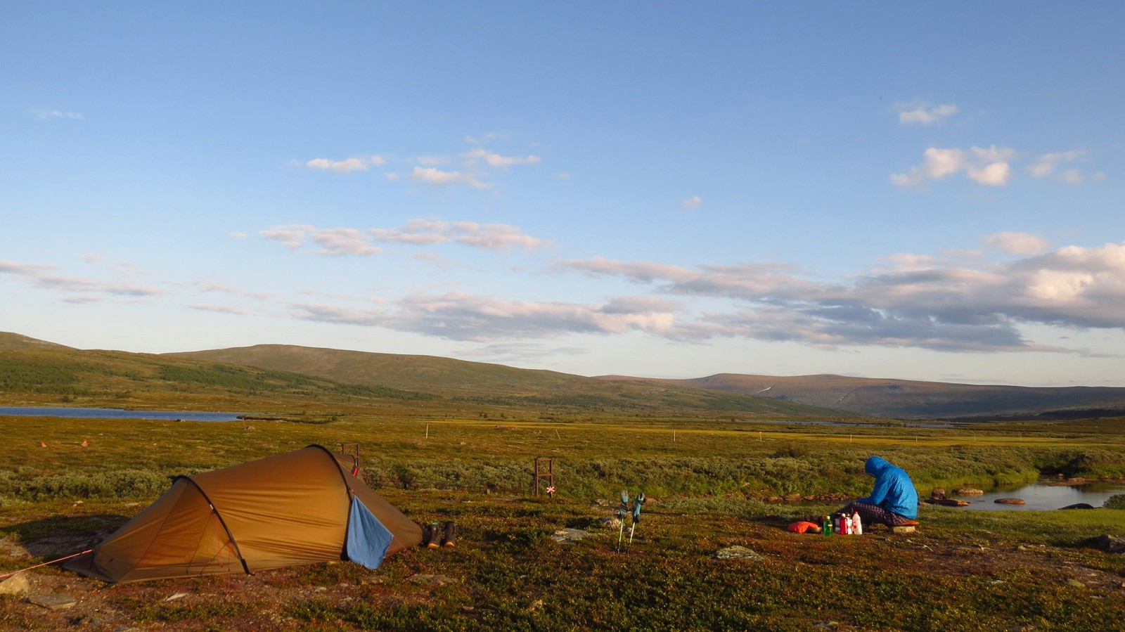 Man with a hooded jacket sits in front of a tent in a landscape in front of a blue sky