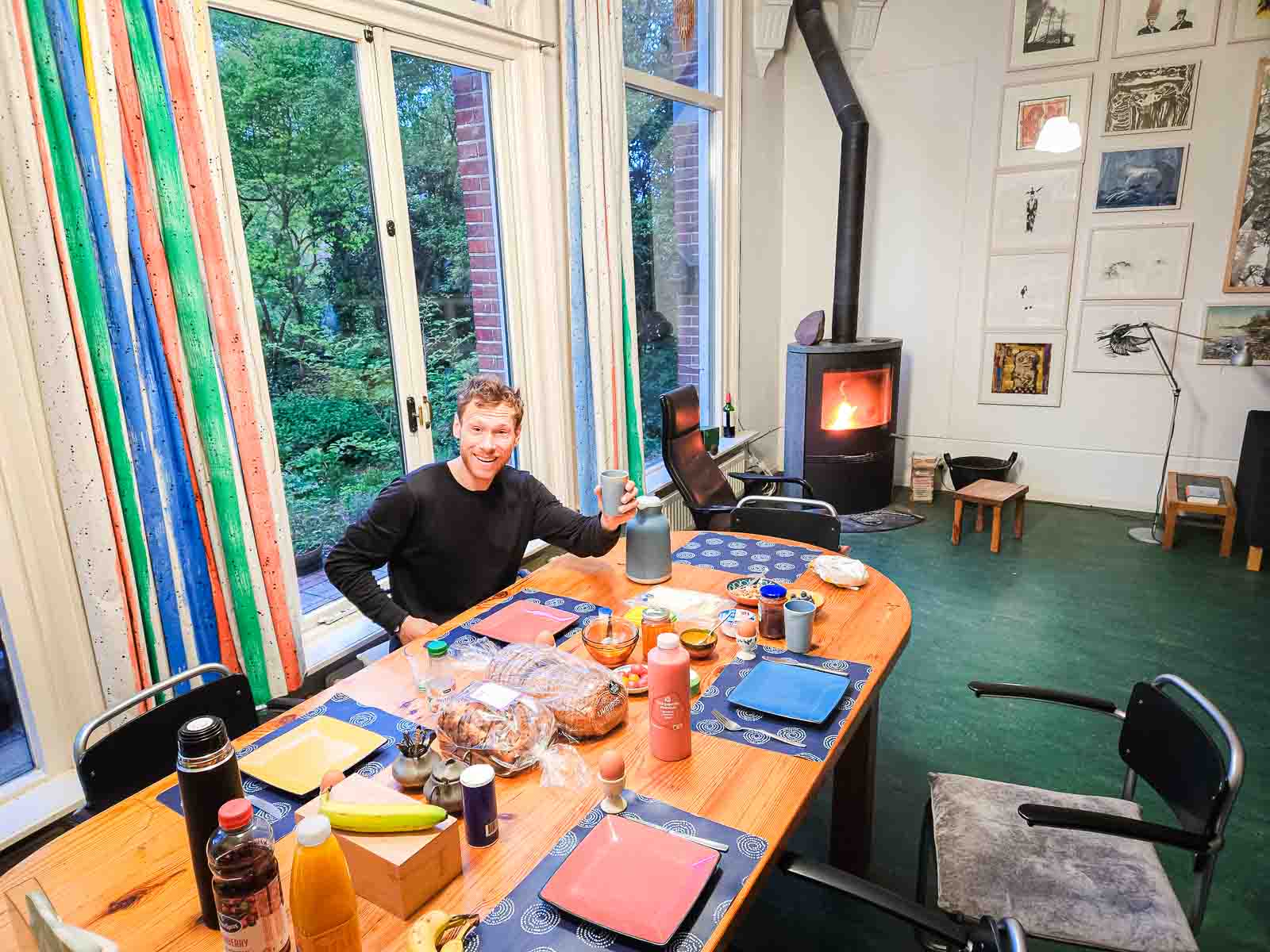 A man sits at a lavishly laid breakfast table and toasts the camera.