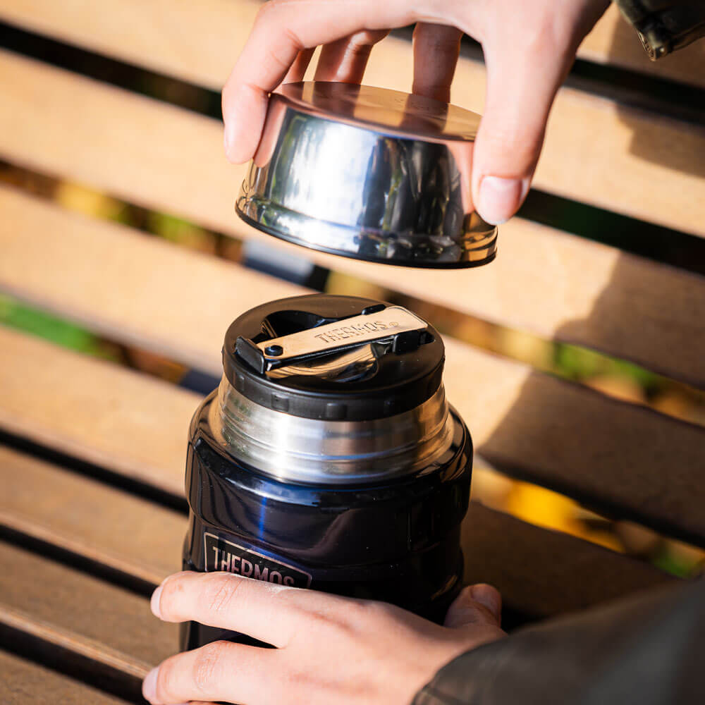 A hand lifts the lid of a thermos flask to reveal the closure underneath. The thermos flask sits on a wooden bench in an autumn setting.
