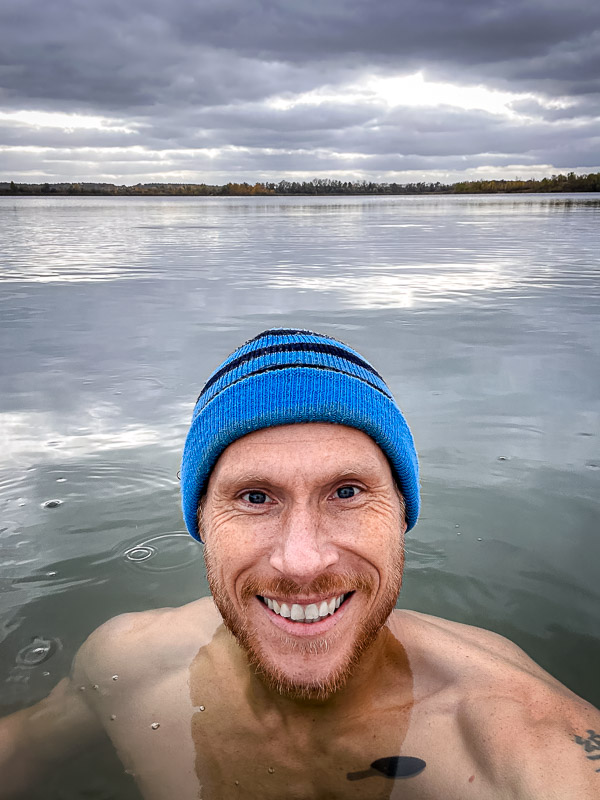 A man wearing a blue beanie smiles at the camera while standing in a cold lake. The sky is cloudy, and the calm water reflects the gray clouds.