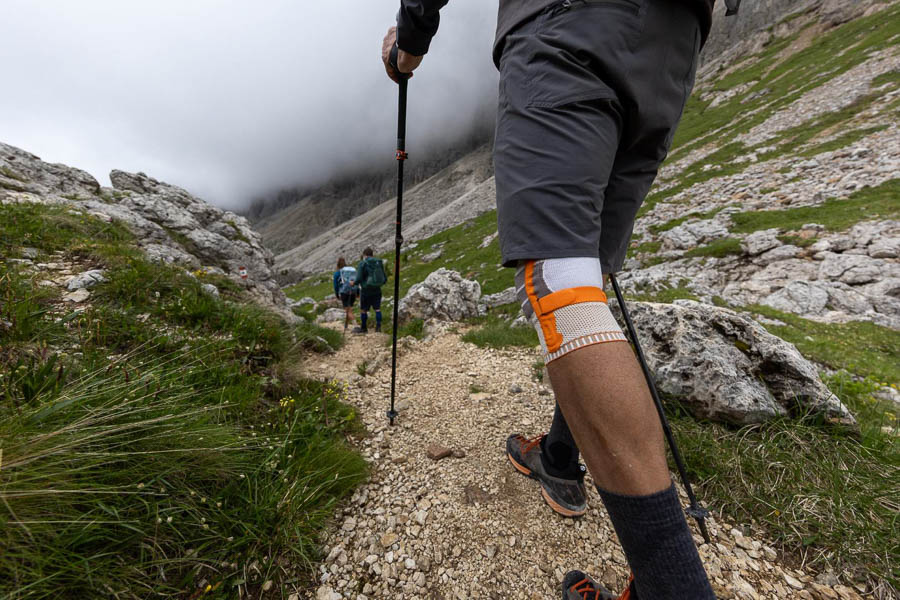 A hiker with trekking poles and a knee brace walks on a rocky trail through a mountainous landscape. Other hikers and clouds over the mountains can be seen in the background.