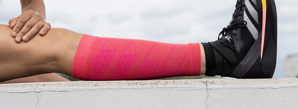 An athlete sits on the ground, stretching their extended leg. They are wearing bright pink compression sleeves on the calf, paired with black running shoes.