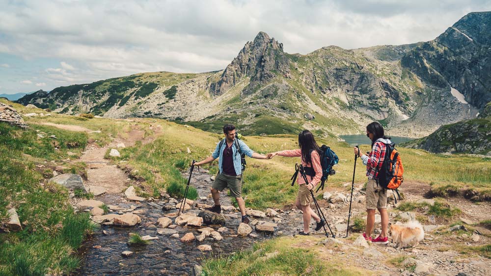 Three hikers, two women and one man, are crossing a mountain stream in an alpine landscape with rocky peaks in the background. The man offers his hand to help – highlighting teamwork and connection to nature.