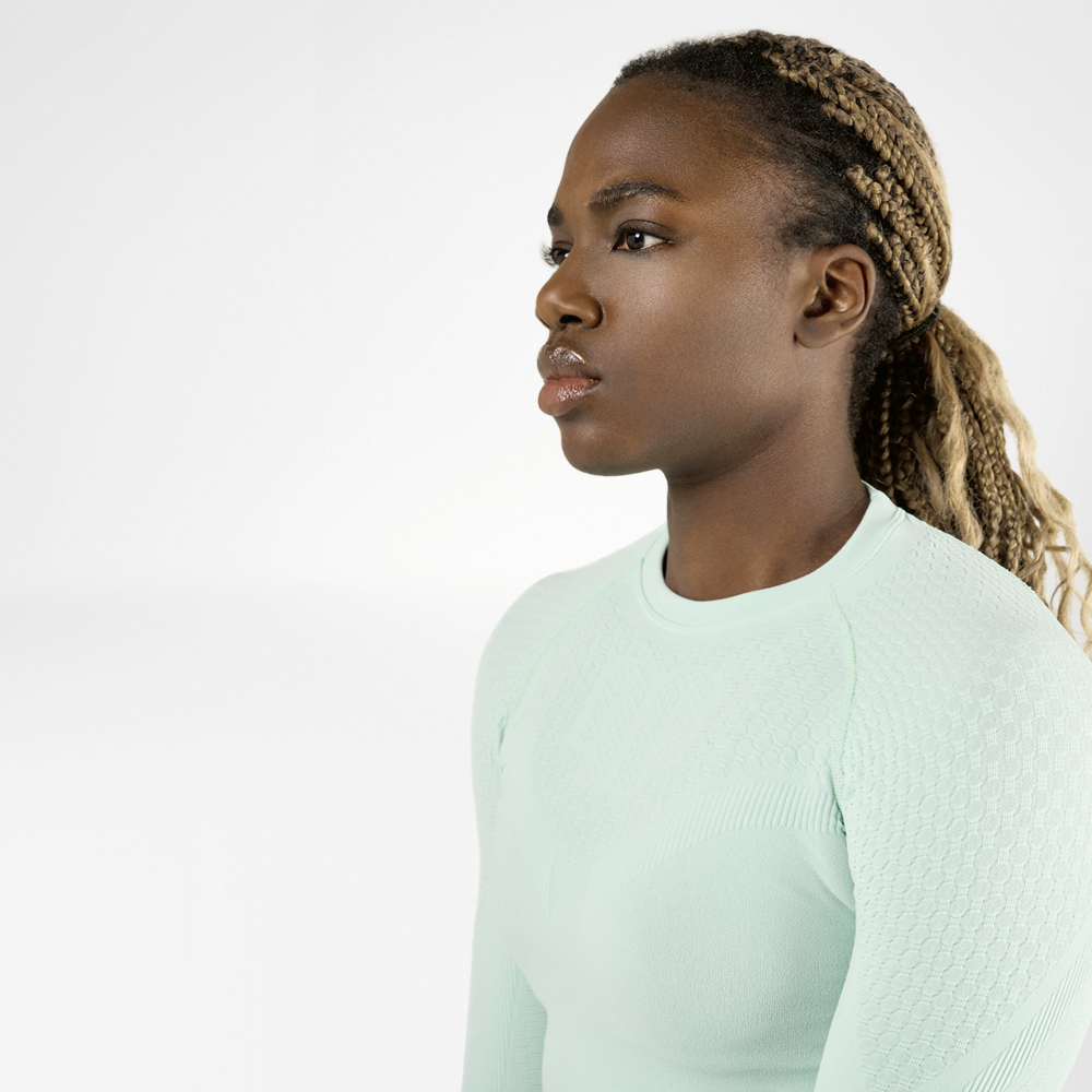 A sporty woman with braided hair wearing a light green, long-sleeved compression shirt is viewed from the side. She has a focused and determined expression on her face.