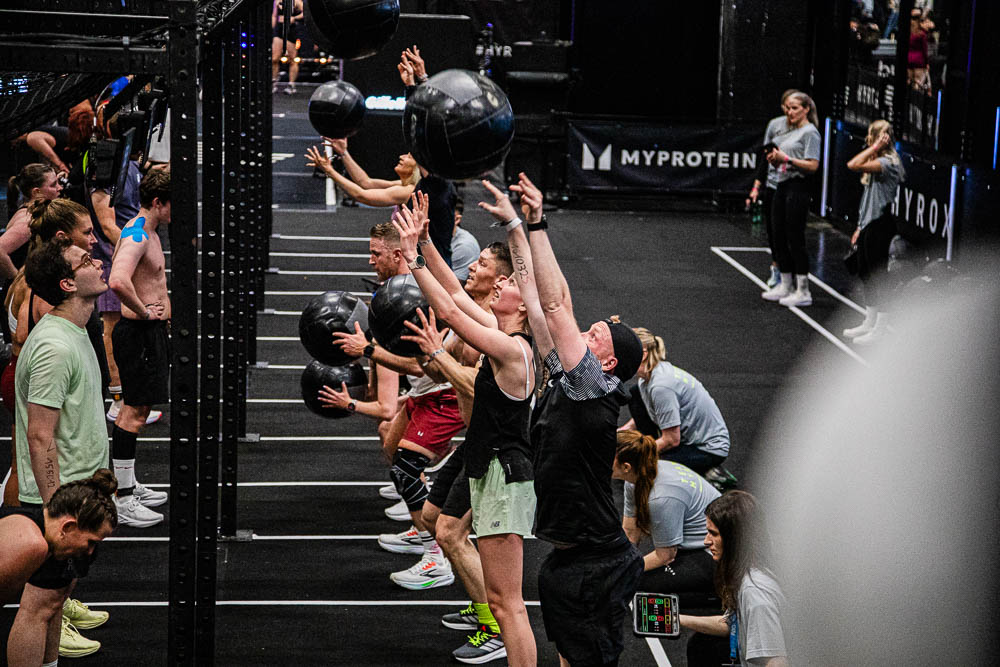 Athletes performing wall balls in a HYROX workout zone, throwing black medicine balls upward while judges and support staff watch the repetitions; the scene shows intense full-body effort, concentration and an energizing competition atmosphere.