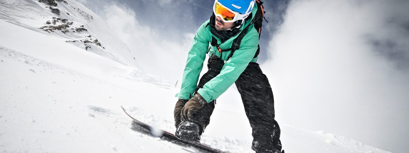 Light frog perspective: Snowboarders in a green jacket attached with his hands a bond and looks down while a lot of snow is whirled around around it