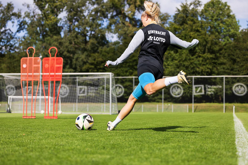 A player from Eintracht Frankfurt takes a powerful shot at the goal in the background. She is wearing training gear with the Eintracht logo and a blue knee support from Bauerfeind Sports.