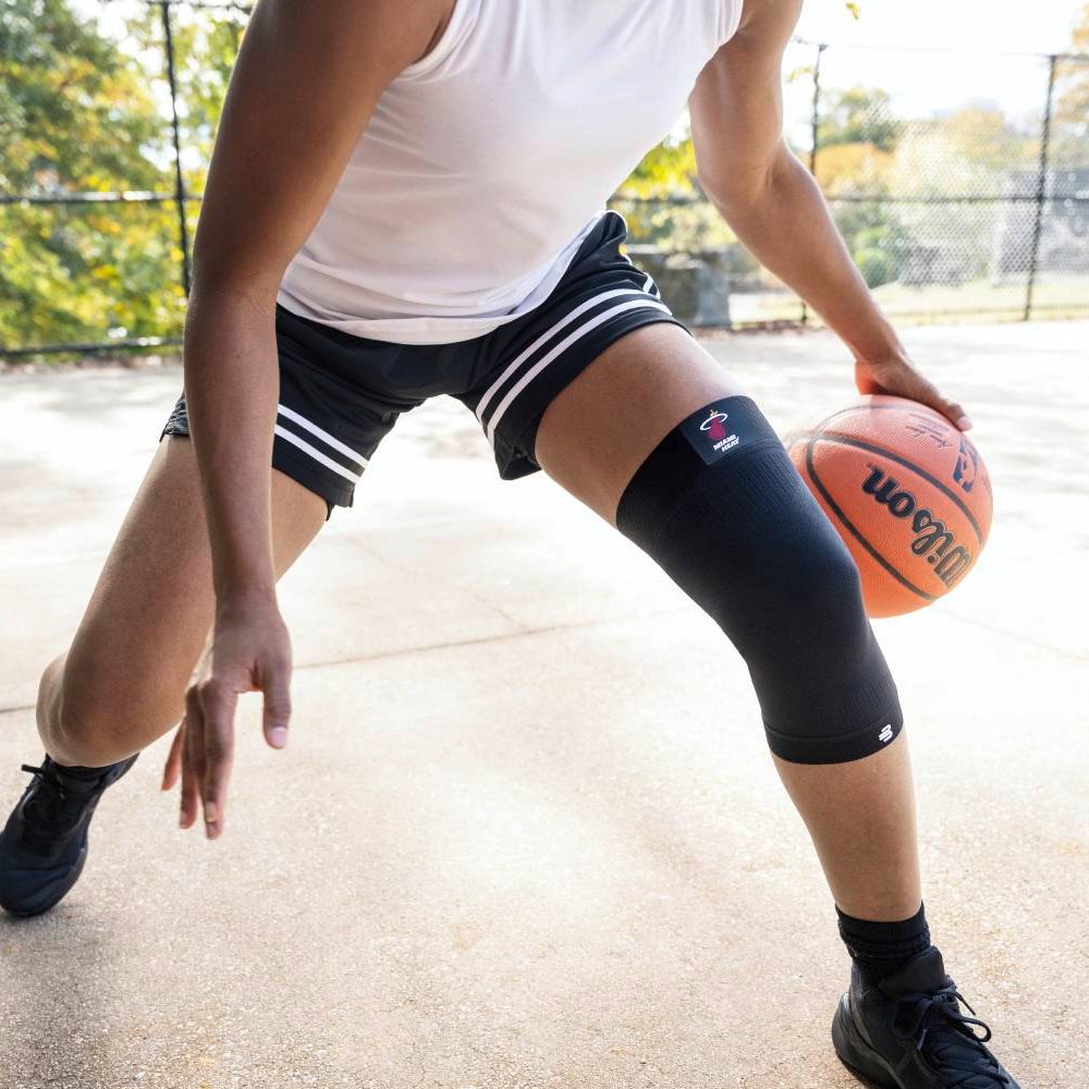 A basketball player wearing a black Miami Heat knee support while defending. Focus on knee support during quick directional changes.