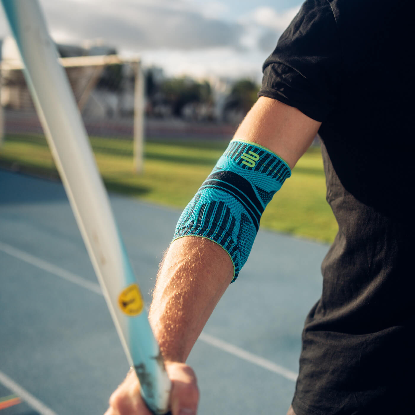 Precision and power – with stable support for your elbow. Close-up of an arm wearing a blue elbow support during a javelin throw. A running track and a sunny landscape are visible in the background.
