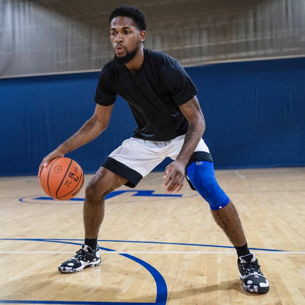 A basketball player in a defensive stance on an indoor court, wearing a blue compression knee sleeve on his right leg. The wooden floor reflects the gym's lighting.