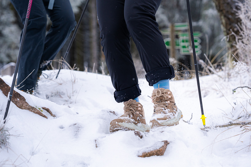 Nahaufnahme von Wanderstiefeln, die durch tiefen Schnee stapfen. Die Füße gehören zu Wanderern mit schwarzen Hosen und blauen Socken. Wanderstöcke mit gelben Spitzen stecken im Schnee, während der Boden von Schnee und Felsen bedeckt ist.