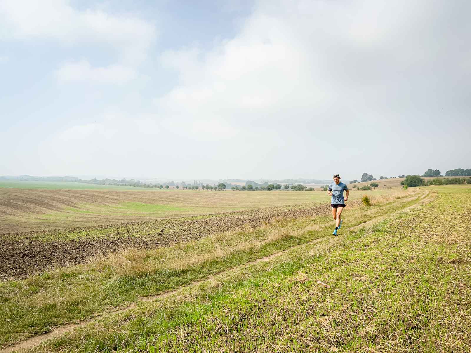 A runner jogging on a rural field path under a slightly cloudy sky. The surroundings are calm and vast, perfect for endurance training.