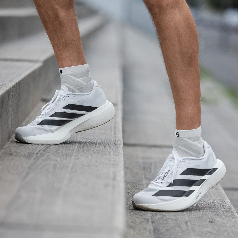 A man stands with slightly bent legs on a staircase. His white short Run Performance Socks and white running shoes with black stripes are visible.