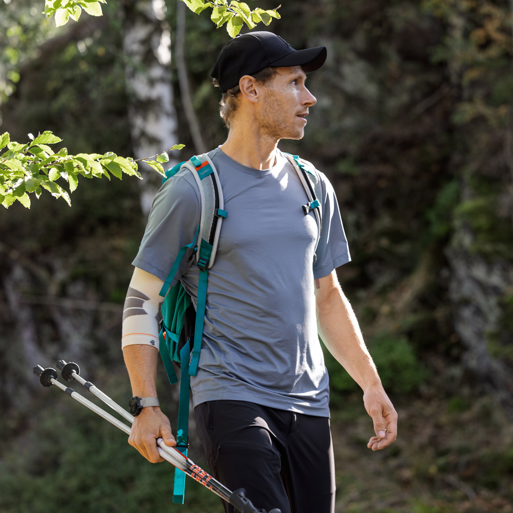 A hiker stands on a shaded forest path, looking attentively into the surroundings. He wears a backpack, holds trekking poles, and uses an Outdoor Compression Elbow Support on his left arm.
