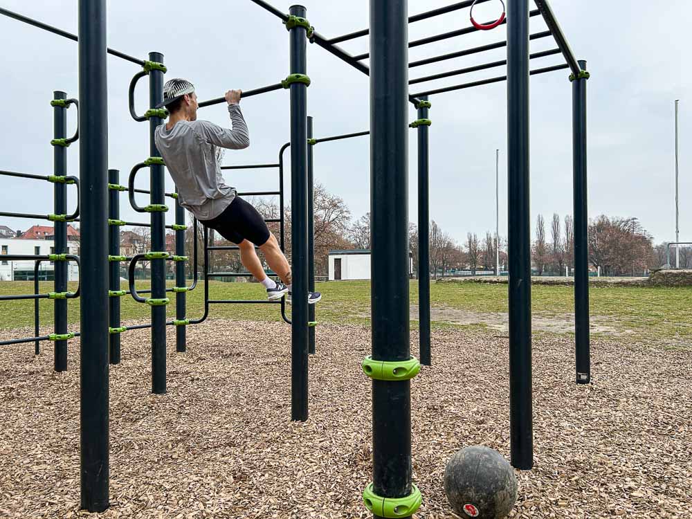 Athlete performs a pulling exercise on an outdoor calisthenics bar during HYROX training; a black training ball lies on the ground.