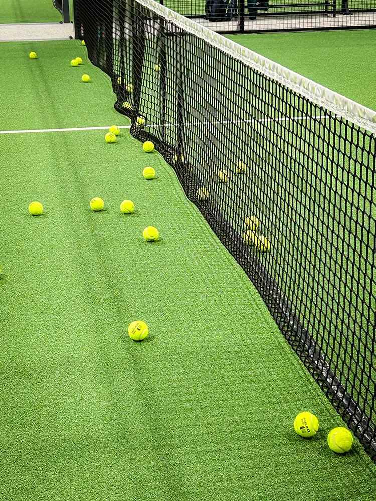 Several yellow padel balls are scattered across the green artificial turf court right next to the net – capturing the vibrant energy after intense rallies.