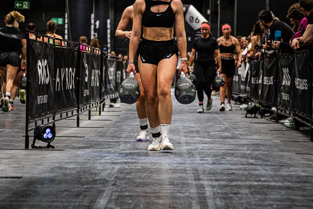 Athlete carrying two heavy kettlebells along the HYROX course during a competition, while other participants follow behind and spectators line the race lane; the scene highlights grip strength