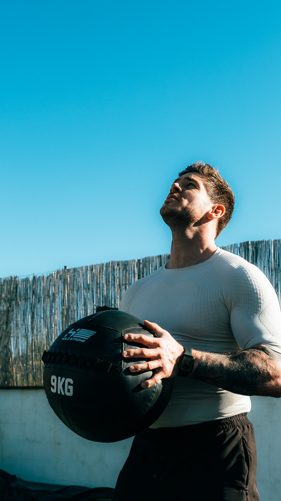 Athlete stands outdoors holding a black 9 kg medicine ball in front of his torso, wearing a light compression shirt and looking upward under a clear blue sky – functional training for hybrid athletes.