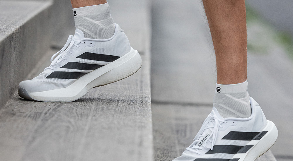 Close-up of white running shoes with black stripes and white compression socks, worn while climbing stairs. The socks extend above the ankle, providing stability and comfort during training.