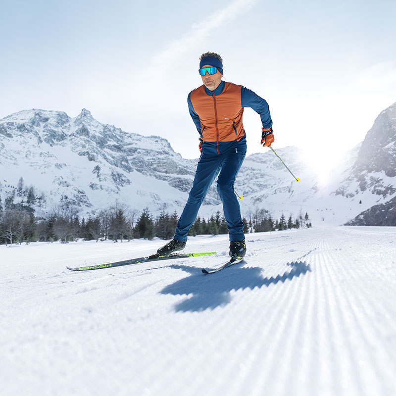 A man with blue ski goggles, photographed from the front, is skiing in a winter landscape. He is wearing an orange jacket and blue ski trousers.