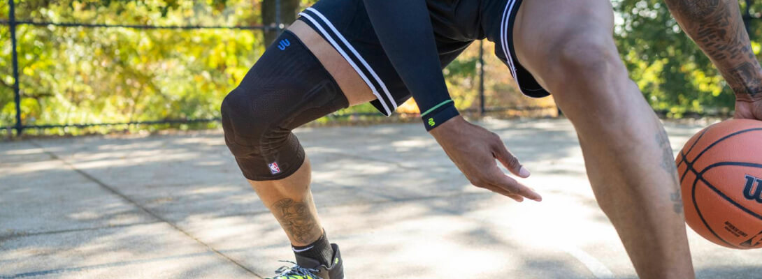 A basketball player wearing a black compression knee support dribbles the ball on an outdoor court. The brace prominently features the NBA logo and manufacturer branding.