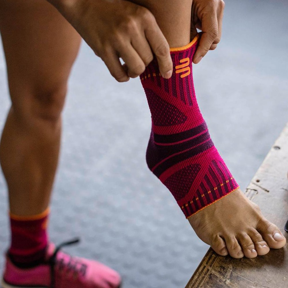 Quick to put on, perfect support! Close-up of a foot with a pink and black ankle support. A hand is pulling the support over the ankle, while a pink sports shoe is visible in the background.