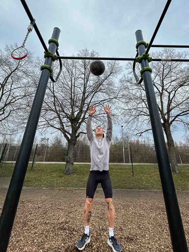 An athlete throws a medicine ball upward in an outdoor training park beneath a calisthenics rig. The scene shows a functional strength and conditioning exercise that fits well into HYROX training without a gym.